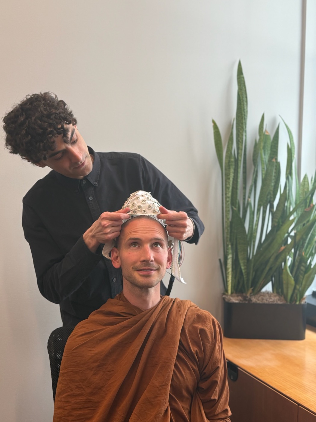 A researcher fits an EEG cap on a smiling monk in robes